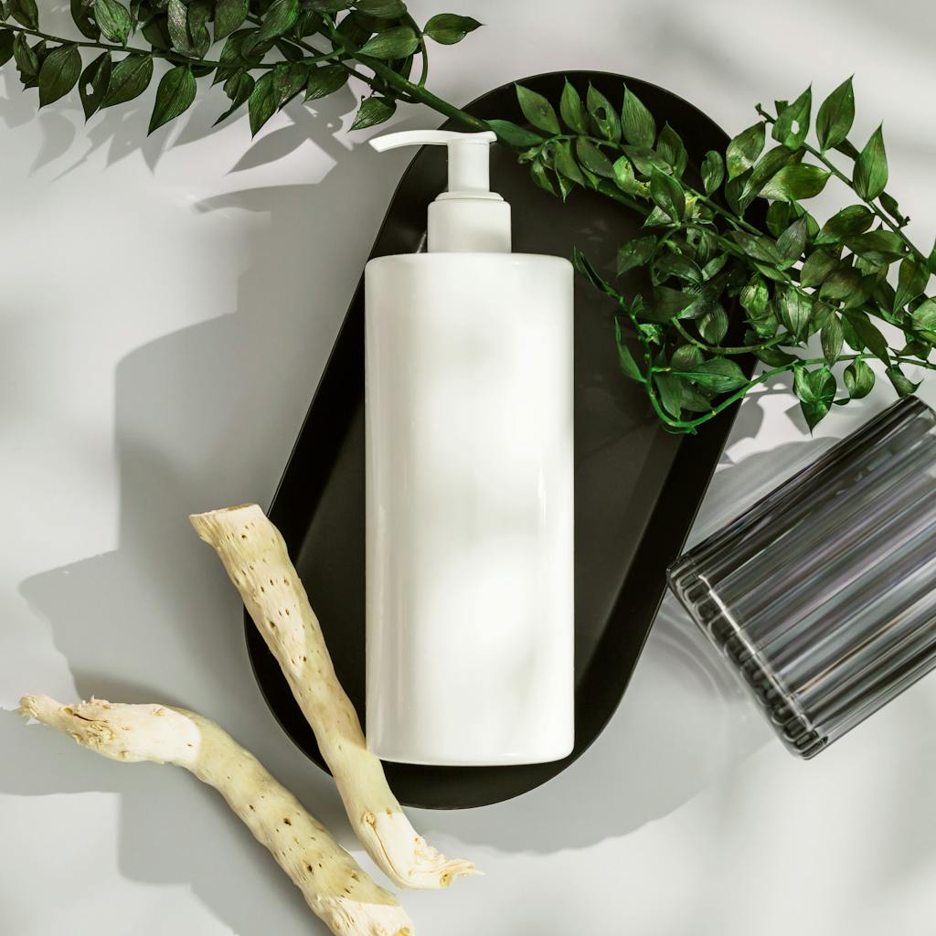 Top view of a white cosmetic bottle with pump surrounded by green leaves and wood on a white surface.