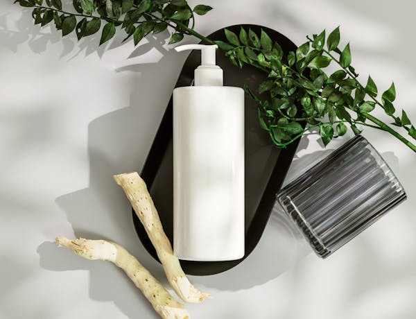 Top view of a white cosmetic bottle with pump surrounded by green leaves and wood on a white surface.
